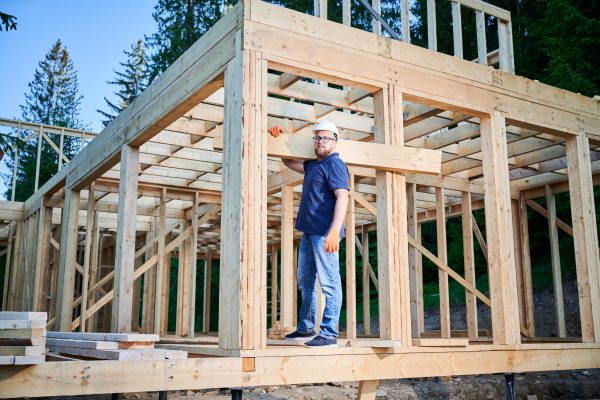 Carpenter constructs wooden-framed house near forest. Man holds large truss on the shoulder while dressed in work clothes and helmet. An idea of modern and eco-friendly construction.